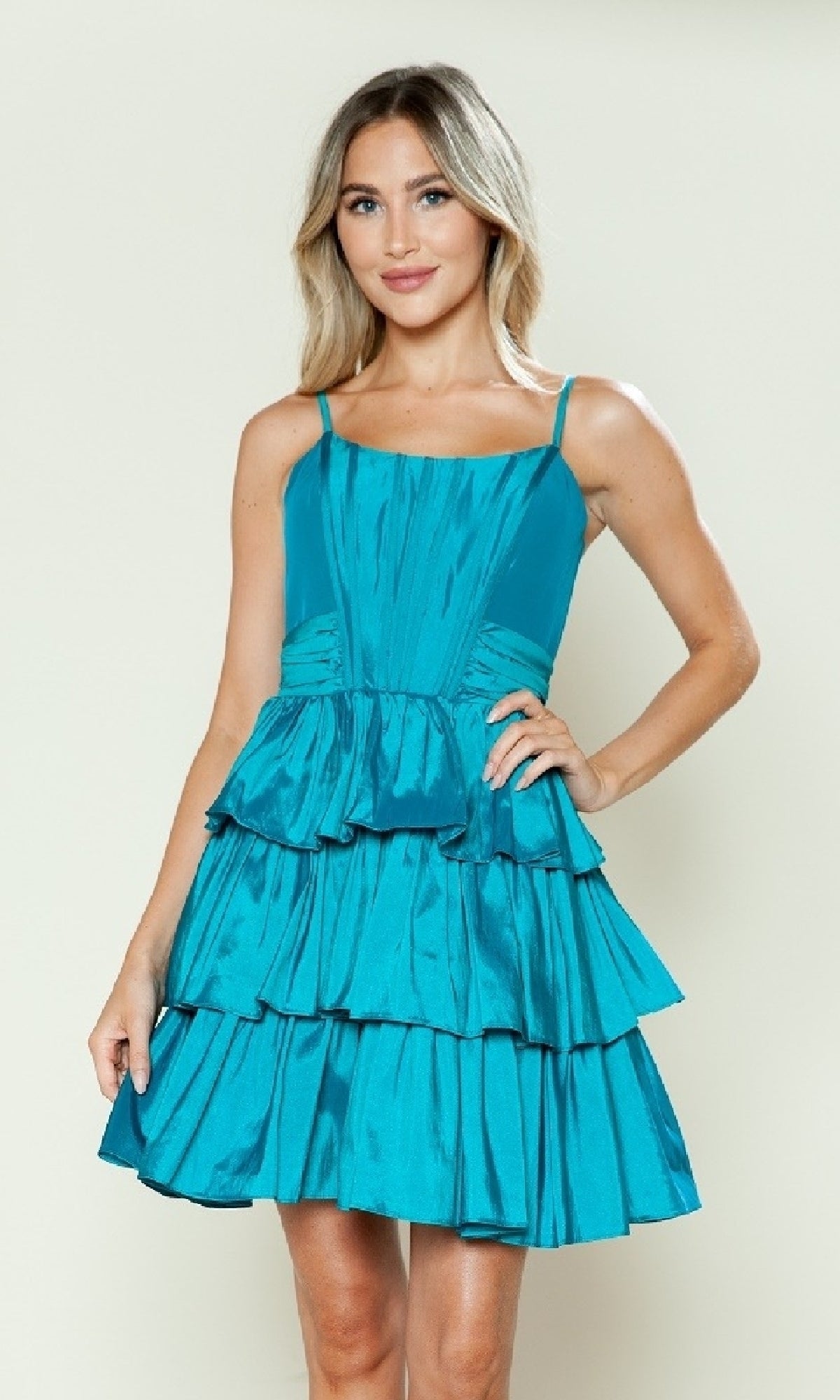 A person models the Short Homecoming Dress: Poly USA 9590, a sleeveless fit-and-flare dress with a teal, tiered A-line skirt, standing against a plain light background facing the camera with one hand on their hip.